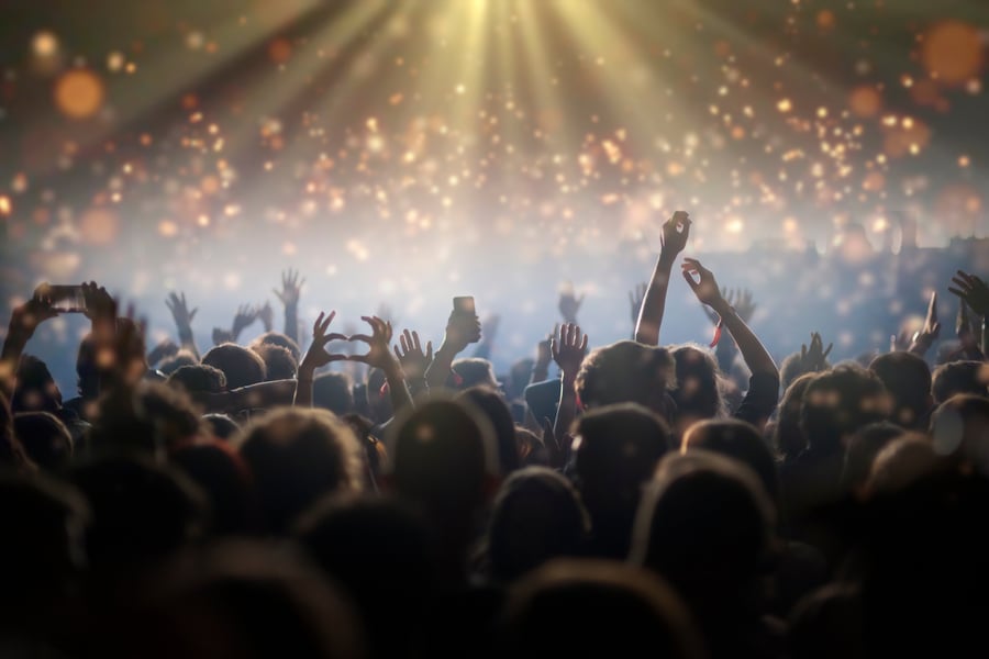 Concert crowd with raised hands and phones, two hands forming a heart, under beams of golden stage light