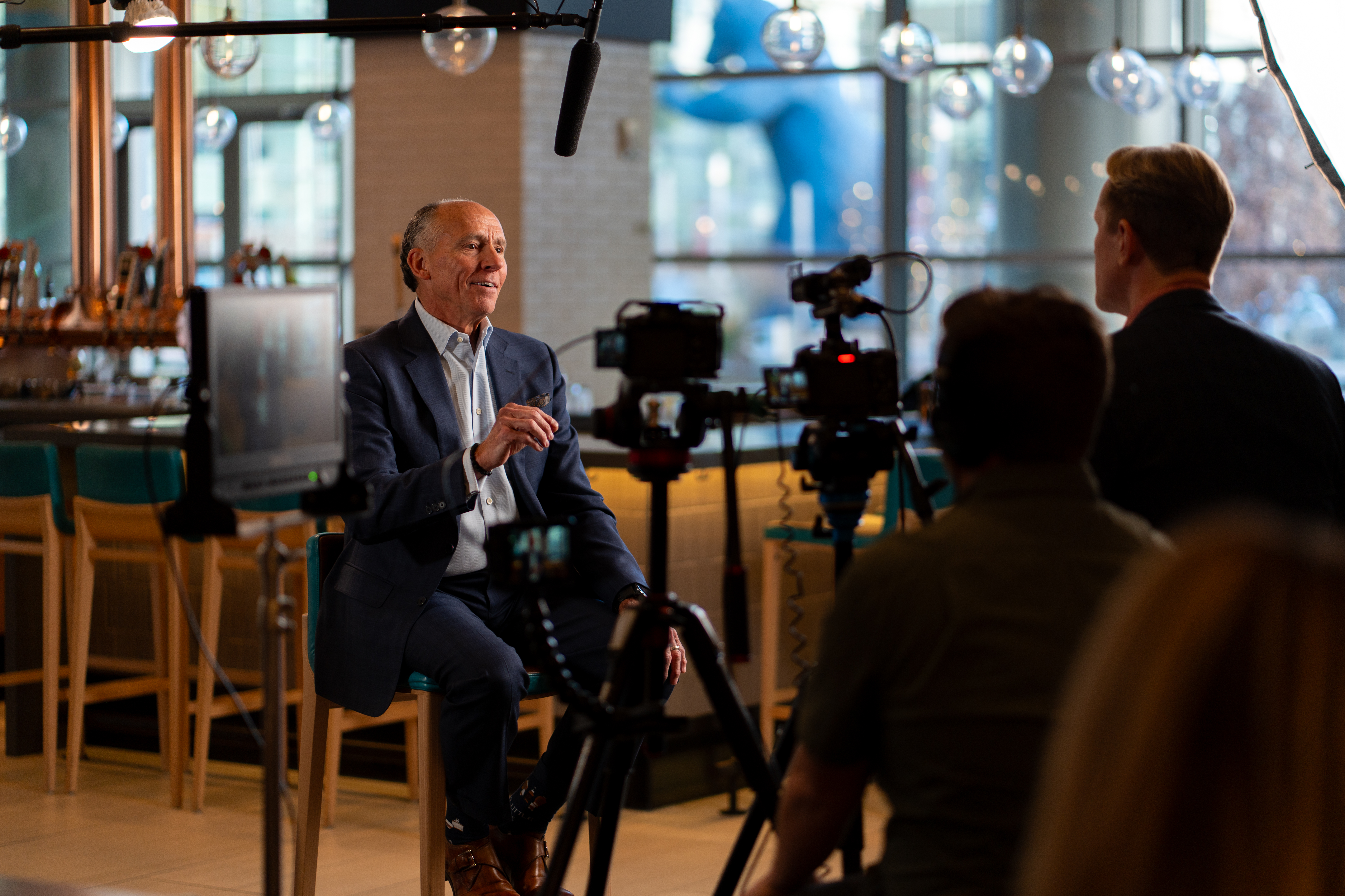 Executive in a navy suit seated for a cinematic sit-down interview, with cameras, a boom mic and crew in the foreground