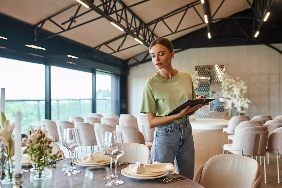 A female event planner holding a clipboard and inspecting place settings at a banquet.