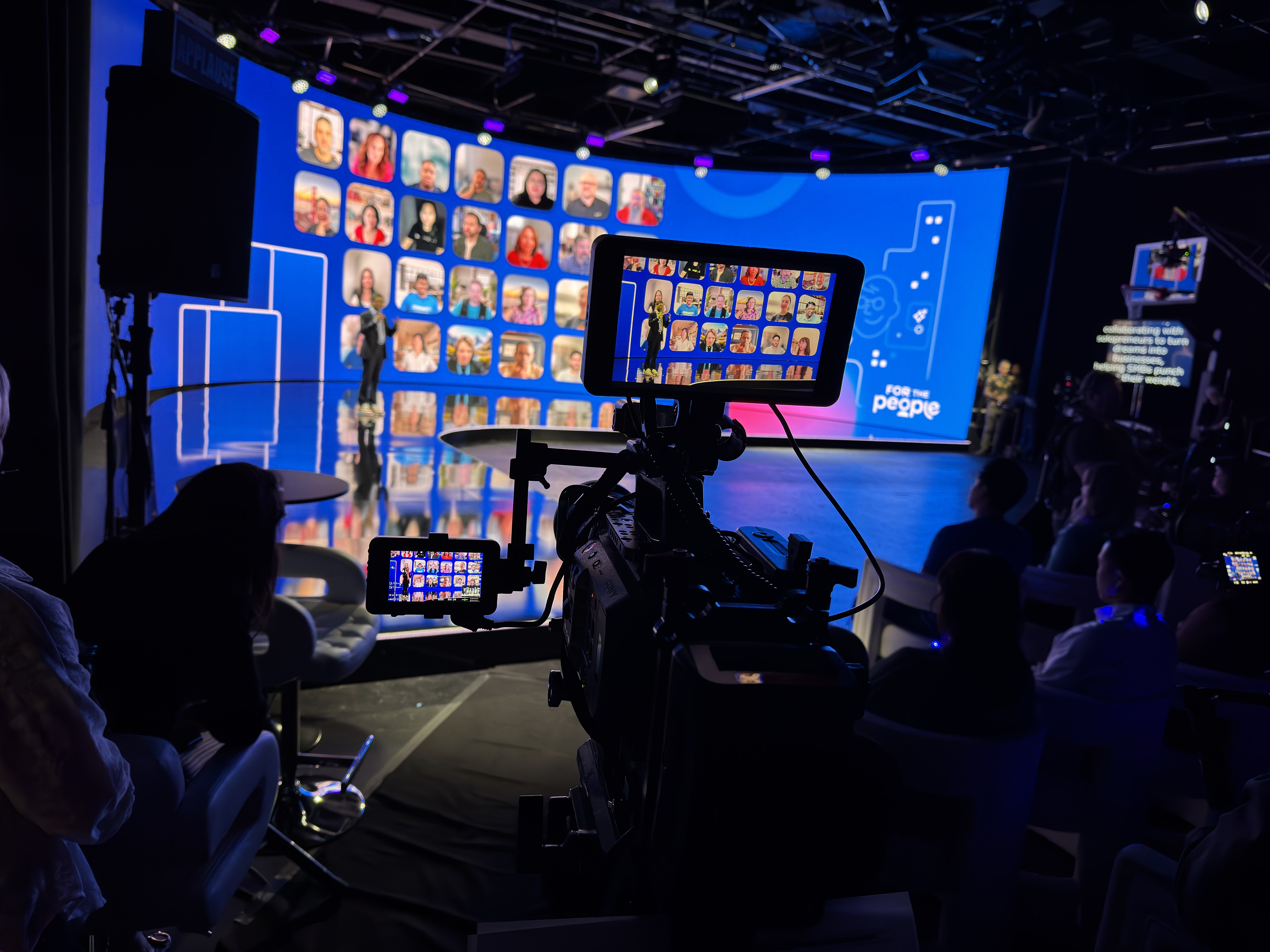 Camera operator’s view of a hybrid event stage, with a presenter in front of a curved LED wall showing a grid of remote attendees on video.